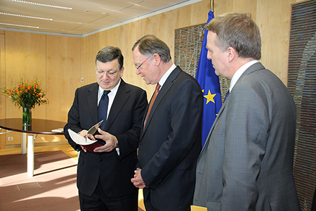 Stehend in einem Büro: von links: Barroso, der sich die überreichten Unterlagen ansieht, Weil und Wenzel