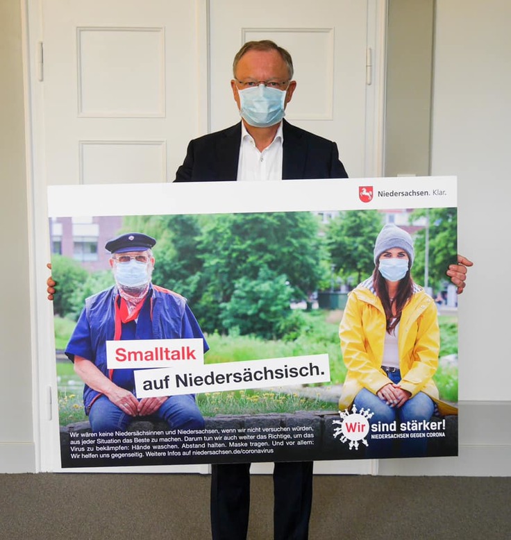 Ministerpräsident Stephan Weil hält ein Plakat, auf dem ein Mann und eine Frau mit einer Mund-Nase-Bedeckung zu sehen sind, die mit deutlich Abstand zueinander schweigend auf einer Bank sitzen. Auf dem Bild steht "Smalltalk auf Niedersächsisch&qu