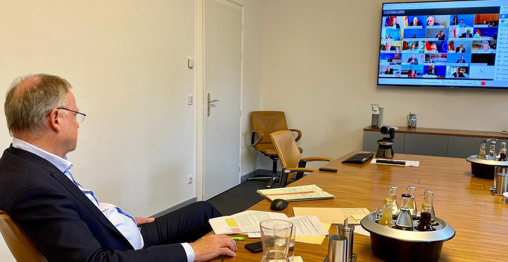 Ministerpräsident Stephan Weil sitzt an einem großen, leeren Konferenztisch, auf dem eien Webcam steht, und blickt auf eine Videokonferenz mit vielen Teilnehmern, die auf einem großen, an der Wand befestigten Bildschirm zu sehen ist.