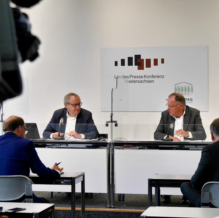 Ministerpräsident Stephan Weil sitzt gemeinsam mit dem Leiter der Landespressekonferenz an einem Tisch vor mehreren Journalisten. Im Hintergrund ist auf einem Schild das Logo und der Aufdruck "Landespressekonferenz Niedersachsen" zu sehen.