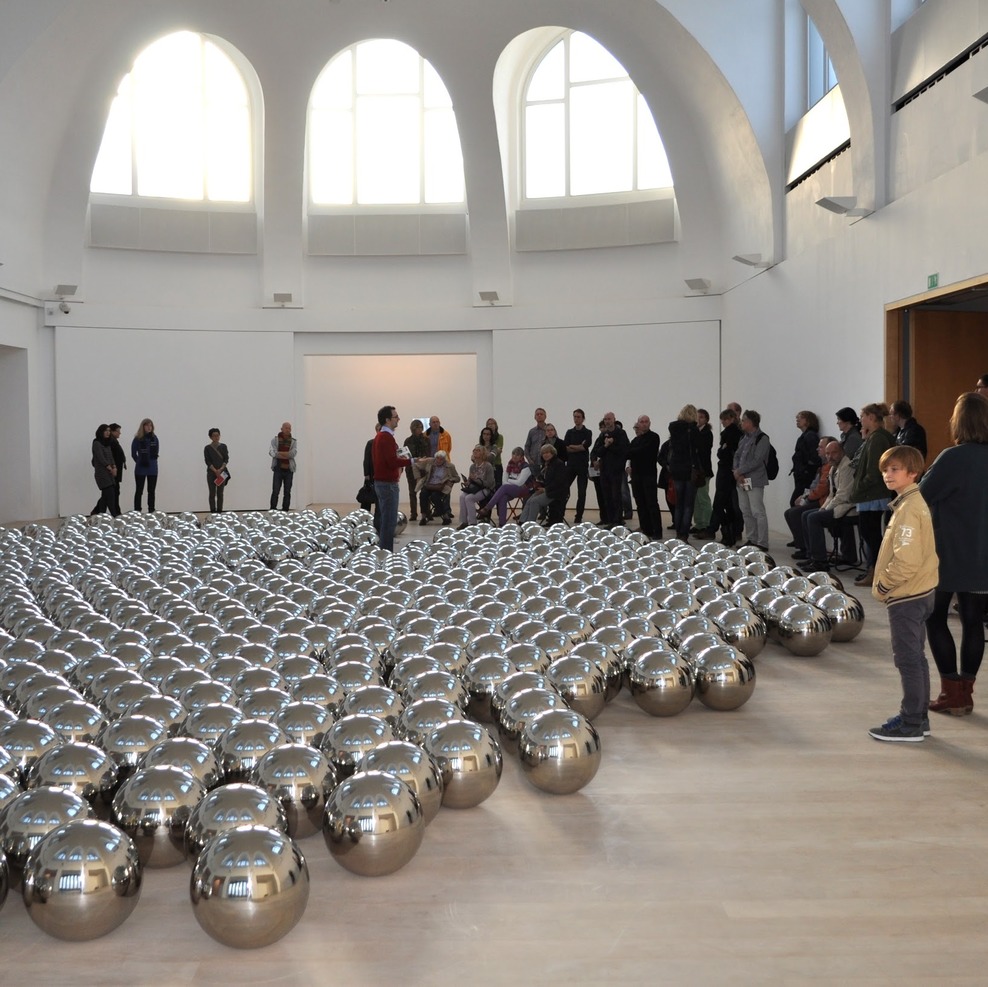 Künstlerische Installation in der kestnergesellschaft in Hannover. Viele silberne Kugeln liegen in einem runden Raum. Besucherinnen und Besucher stehen im Halbkreis daneben und betrachten sie.