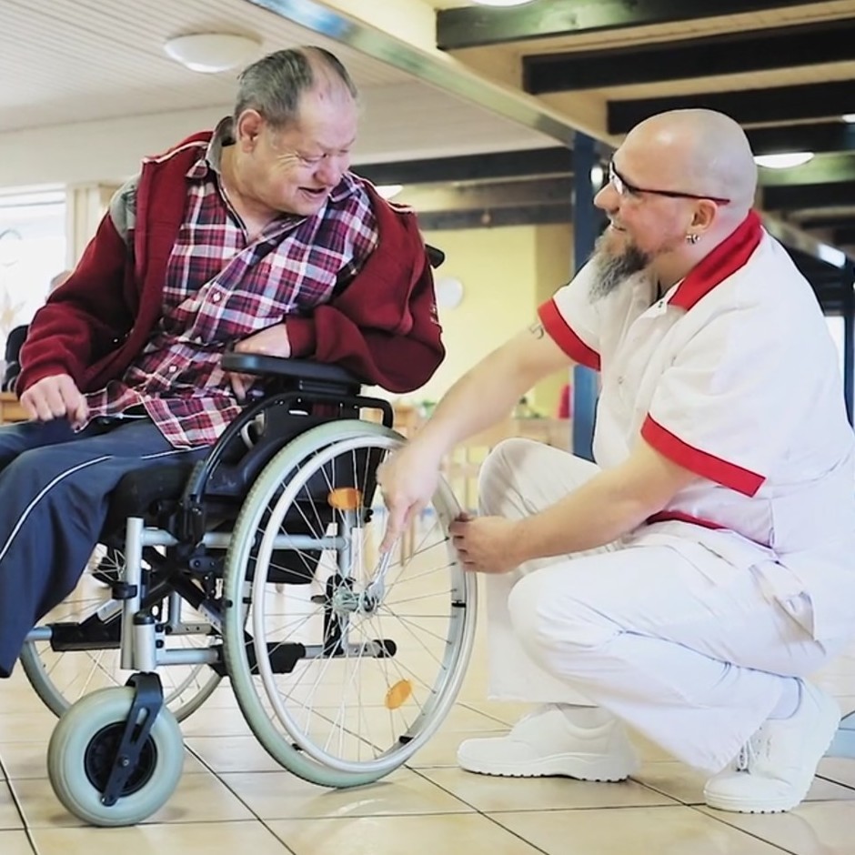 Screenshot aus dem Kinospot "Ich pflege gern": Ein Pfleger hilft einem Bewohner mit dessen Rollstuhl.