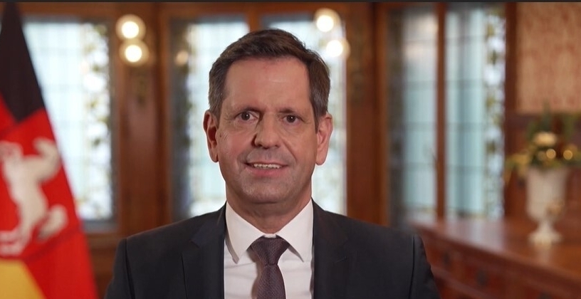 Ministerpräsident Olaf Lies steht im Gästehaus der Niedersächsischen Landesregierung, hinter ihm die Landesflagge Niedersachsens.