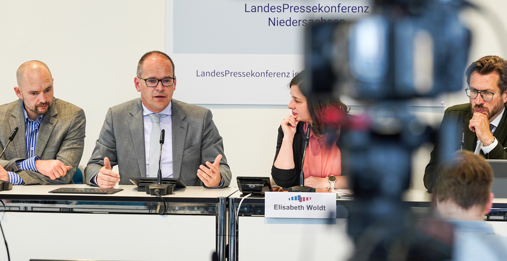 Blick auf ein Podium mit drei Männern und einer Frau, einer der Männer spricht zum Publikum. An der rechten Seite zeichnet sich unscharf eine Kamera ab.